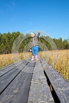 Little child with arm up and narrowing eyes standing on background on a wooden planks pathway, swamp land