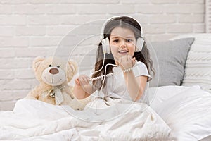 Little girl listening to the music with the headphones on bed.