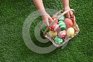 Little child with basket of painted Easter eggs on green grass, top view.