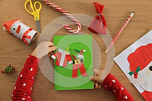 Little child with beautiful Christmas greeting card at wooden table, top view