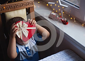 Little child boy hiding behind red heart shaped gift box with bow near window in daylight