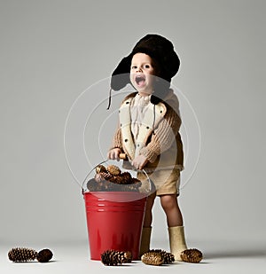 Little child boy in winter retro hat stand and hold in hand red Christmas bucket full of fir cones