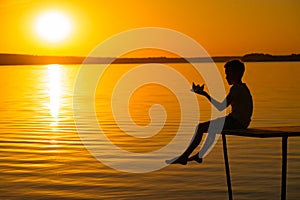 A little child is on a bridge with origami in the shape of a boat in his hands at sunset. The legs of the child lowered