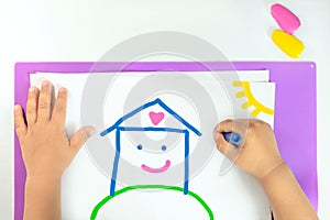 little child draws a house with wax crayons on white paper, concept of a happy childhood and family, home sweet home