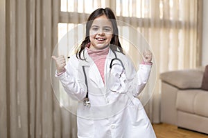 Little child dressed in doctor`s uniform giving thumbs up in a fun and positive attitude. She is at home