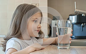 Little child is drinking clean water at home, close up. Caucasian cute girl with long hair is holding a water glass in her hand