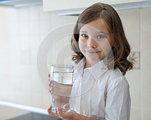 Little child is drinking clean water at home, close up. Caucasian cute girl with long hair is holding a water glass in her hand