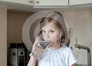 Little child is drinking clean water at home, close up. Caucasian cute girl with long hair is holding a water glass in her hands