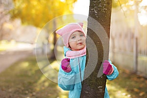 Little child girl 4 years in blue jacket, red pants, fur hat smiles and peeps fervently from behind tree, dried twig in fingers on