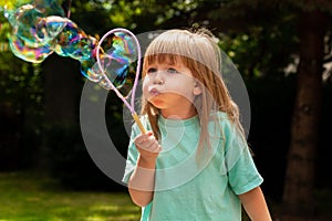 Little child, girl blowing huge bubbles alone, portrait outdoors. Young kid with puffed cheeks blows big bubbles outside, closeup