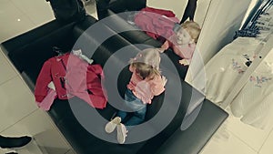 Little child girl in a clothing store sits on the couch in front of a mirror