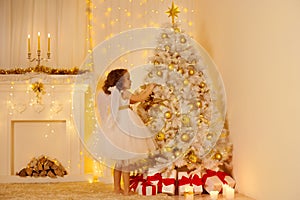 Little Child Girl Decorating Christmas Tree with Golden Ball in Cozy White Interior with Fireplace. Xmas Room Lights