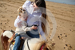 Little child girl feeling worried and hugging her mother while riding pony on the sandy beach