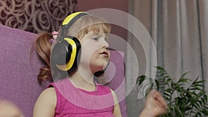 Little child girl in headphones enjoying listen music. Dancing on sofa at home