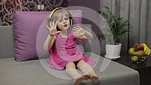 Little child girl in headphones enjoying listen music. Dancing on sofa at home