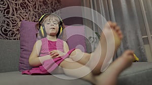 Little child girl in headphones enjoying listen music. Dancing on sofa at home