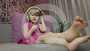 Little child girl in headphones enjoying listen music. Dancing on sofa at home