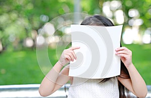 Little child girl hiding face behind blank white paper in the green garden
