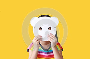 Little child girl holding blank white animal paper mask fronting her face isolated on yellow background. Idea and concept for kid