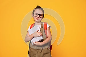 Little child girl holding book, smiling looking at camera, dressed in casual clothes and trendy glasses. Back to school