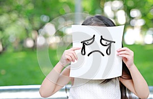 Little child girl holding up a white paper with draw crying face expression in nature park