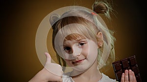 Little child girl kid eating chocolate bar dessert, showing thumbs up isolated on dark background