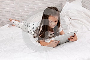 Little child girl lies in bed uses digital tablet.