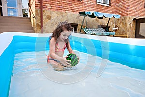 Little child girl plays with a floating ball while having fun in the inflatable water pool at home backyard on sunny day