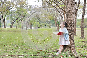 Little child girl reading book in summer park outdoor standing lean against tree trunk with looking to sky