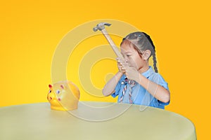 Little child girl in school uniform taking hammer trying to broke piggy bank isolated on yellow background at table. Schoolgirl