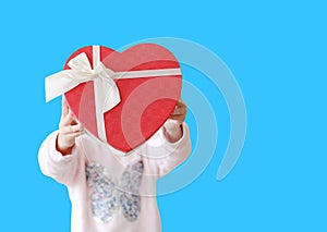 Little child girl showing red heart gift box  on blue background. Kid giving red heart gift box for you with copy space.