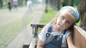 Little child girl sitting alone on a bench in summer park.