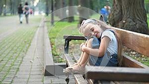 Little child girl sitting alone on a bench in summer park.