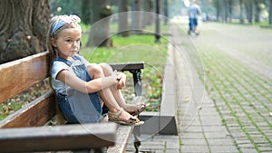 Little child girl sitting alone on a bench in summer park