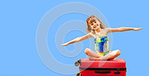 Little child girl wear swimsuit sitting on suitcase isolated on blue background with hands up, show plane. Vacation concept