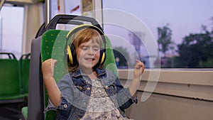 Little child girl wearing headphones listening music, relax dancing while traveling by bus in city