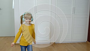 Little child girl wearing headphones listening to music and funnily dancing near wardrobe at home