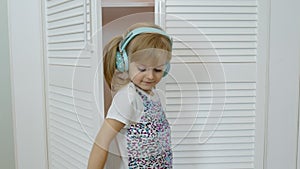 Little child girl wearing headphones listening to music and funnily dancing near wardrobe at home