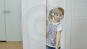 Little child girl wearing headphones listening to music and funnily dancing near wardrobe at home