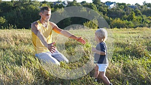 Little child goes on green grass at the field to his father at sunny day. Dad lifting up his baby boy at nature. Happy