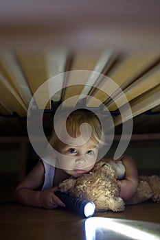 Little child, hiding under the bed, hugging teddy bear and holding flashlight