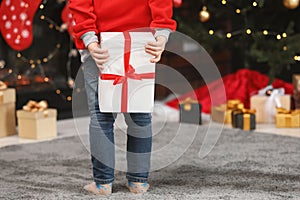 Little child holding gift box behind his back at home, closeup. Christmas celebration