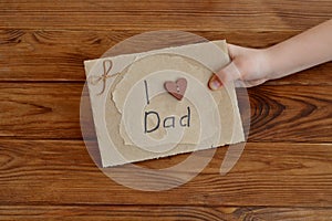 Little child holds a card I love dad. Postcard made of cardboard and wrapping paper, decorated with wooden heart, waxed cord