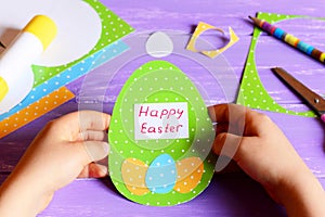 Little child holds a Easter paper card in hands. Child shows Easter greeting card in egg shape. Materials for kids activity