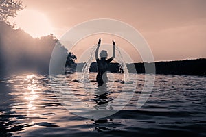 Little child jumping and splashing water in rural pond in monochrome sunset