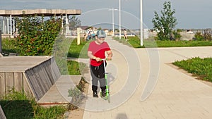 Little child learning to ride a scooter in a city park on sunny summer day. Cute preschooler boy in safety helmet riding