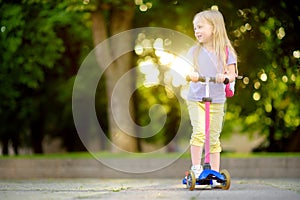 Little child learning to ride a scooter in a city park on sunny summer day. Cute preschooler girl in safety helmet riding a roller