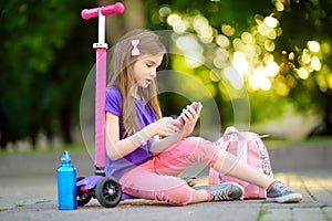 Little child learning to ride a scooter in a city park on sunny summer day. Cute preschooler girl in safety helmet riding a roller