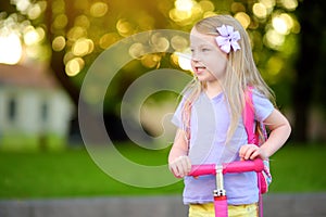 Little child learning to ride a scooter in a city park on sunny summer day. Cute preschooler girl in safety helmet riding a roller