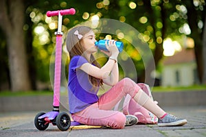 Little child learning to ride a scooter in a city park on sunny summer day. Cute preschooler girl in safety helmet riding a roller